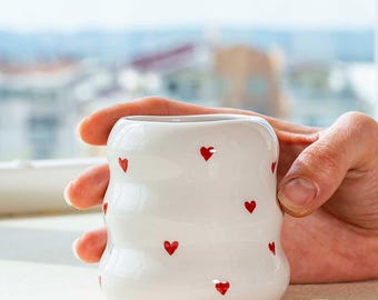 May include: White ceramic mug with a wavy design, adorned with small red heart shapes. The mug is held in a hand, showcasing its unique form and decorative pattern. A charming and simple design.