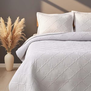 May include: White quilted bedspread and two matching pillowcases with a geometric design. The bedding is arranged on a bed. A vase with dried pampas grass is on the left side of the bed.