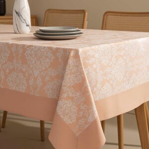 May include: A peach-colored tablecloth with a white floral pattern covers a wooden table. The tablecloth has a solid peach border. Three gray plates are stacked on the table. A vase with dried flowers is in the background.