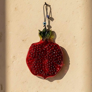 Könnte beinhalten: Ein rotes Glasgranatapfel-Ornament mit grün-gelber Spitze, das an einer schwarzen Schnur hängt. Das Ornament hat eine strukturierte Oberfläche und einen kleinen blauen Augenanhänger. Der Hintergrund ist eine hellbeige Wand.