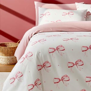 Könnte beinhalten: Weißes Bettwäsche-Set mit einem Muster aus rosa Schleifen. Das Set beinhaltet einen Bettbezug und Kissenbezüge. Der Hintergrund ist einfarbig rosa. Ein Weidenkorb befindet sich im Hintergrund.