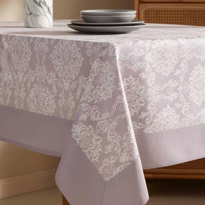 May include: A light purple tablecloth with a white floral pattern and a solid purple border. The table is set with gray plates and a decorative marble vase. The tablecloth is made of a soft, durable fabric.