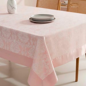 May include: A pink tablecloth with a white floral pattern and a solid pink border. Two gray plates are stacked on the table. A marble vase with dried plants sits on the table. Two wicker chairs are in the background.