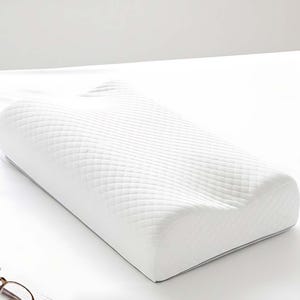 May include: White memory foam pillow with a quilted pattern. The pillow has a curved design for neck support and a gray trim. The pillow is on a white surface, with a book and glasses in the background.