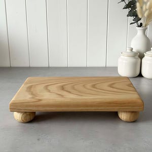 Puede incluir: Una tabla de servir rectangular de madera clara con esquinas redondeadas y cuatro pequeños pies redondeados. La veta de la madera es visible. La tabla está sobre una superficie gris, con jarrones y tarros blancos en el fondo. Un artículo decorativo para la cocina.