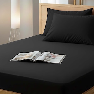 Peut inclure: Un ensemble de lit noir comprenant un drap-housse, deux taies d'oreiller et un magazine ouvert. Le cadre de lit est en bois clair. Un lampadaire blanc est positionné à gauche du lit.