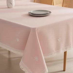 May include: A light pink tablecloth with white floral embroidery and a fringed edge. Two gray plates sit on the table. The table is set with a vase and two chairs in the background.