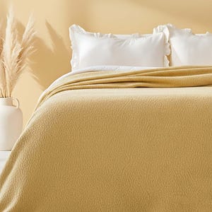 May include: A mustard-yellow blanket covers a bed with white ruffled pillowcases. The blanket has a textured surface and drapes over the edge of the bed. A white vase with dried pampas grass sits to the left.