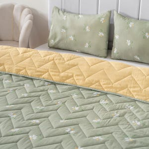 Peut inclure: Une couette vert sauge et deux taies d'oreiller assorties avec un motif floral de marguerites blanches et une texture piquée en chevron. La couette est repliée pour révéler un envers jaune pâle uni avec un motif piqué en chevron assorti.