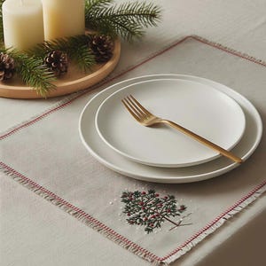May include: A festive table setting with a cream-colored tablecloth featuring a red border and an embroidered tree design. Includes white plates, a gold fork, and candles with pine and pinecones.