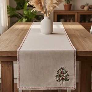 May include: A cream-colored table runner with a red stitched border and a Christmas tree design. The tree is embroidered with green needles and red berries. A white vase with dried pampas grass sits on the runner.