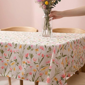 May include: A floral tablecloth with a pattern of pink and yellow flowers and green leaves. The tablecloth is on a wooden table with a vase of flowers. The flowers in the vase are pink, yellow, and purple.