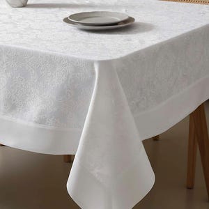 May include: A white tablecloth with a subtle floral pattern covers a wooden table. The tablecloth has a wide, plain border. Two gray plates are on the table. The scene suggests a dining setting.