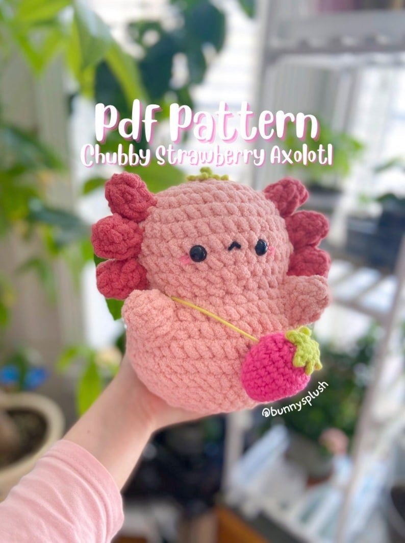 Puede incluir: Un patr&oacute;n de crochet para un juguete de peluche axolotl fresa rosa y verde. El patr&oacute;n est&aacute; disponible para descargar en PDF. La imagen muestra un juguete de peluche terminado que est&aacute; siendo sostenido por una mano.
