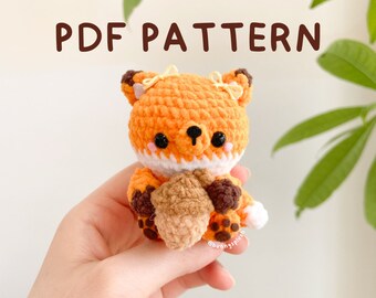 May include: A crocheted fox amigurumi toy holding a brown acorn. The fox is orange with white accents and has black eyes. The toy is about 6 inches tall and is holding the acorn in its paws.