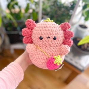 Puede incluir: Un juguete de peluche axolotl de crochet rosa con una hoja verde en la cabeza y una fresa de crochet rosa colgando de su cuello. El juguete est&aacute; siendo sostenido por una mano. El texto "@bunnyplush" es visible en la fresa.