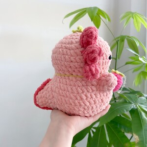 Puede incluir: Un juguete de peluche axolotl de crochet rosa con una banda el&aacute;stica amarilla alrededor del cuello. El axolotl tiene detalles rosas y verdes y est&aacute; siendo sostenido por una mano.