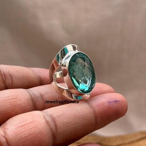 Op de afbeelding: Een zilveren ring met een grote, ovale, lichtblauwe edelsteen. De ring heeft een modern ontwerp met een brede band en een bezel setting. De tekst "JewelrygiftIndia" is zichtbaar.