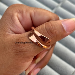 May include: A copper-colored ring with a unique, open design. The ring wraps around the finger with two curved, elongated shapes. The text "JewelrygiftIndia" is visible in the image.