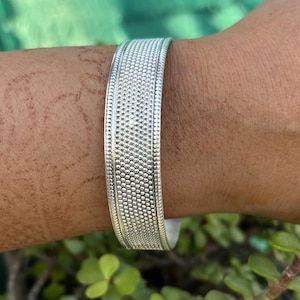 May include: A silver bangle bracelet with a textured design. The bracelet is made of a smooth, shiny metal and has a simple, elegant look.