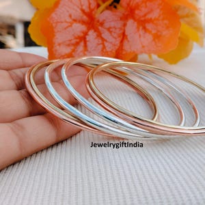 May include: A selection of thin bangles in silver, rose gold, and yellow gold. The bangles are stacked, displaying their different colours and textures. The image is set against a soft, neutral background. The text "Jewelrygiftindia" is visible.