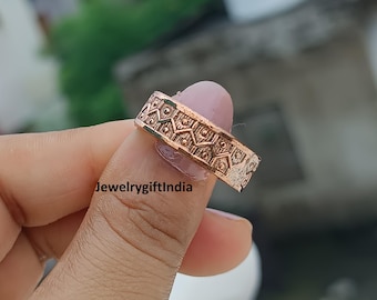 Op de afbeelding: Een koperkleurige ring met een geometrisch patroon. De ring wordt tussen de vingers gehouden en toont het ingewikkelde ontwerp van herhalende vormen en lijnen. Het oppervlak van de ring heeft een textuur en de algehele stijl is vintage.