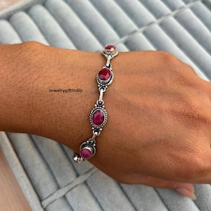 May include: A silver bracelet with oval, deep red gemstones. The bracelet has an ornate design with a rope-like border around each stone. The text "JewelrygiftIndia" is visible on the left side of the image.