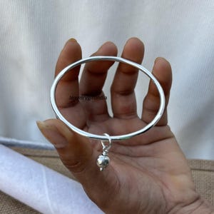 Könnte beinhalten: Ein silbernes Armreif-Armband mit einem kleinen silbernen Kugel-Anhänger.