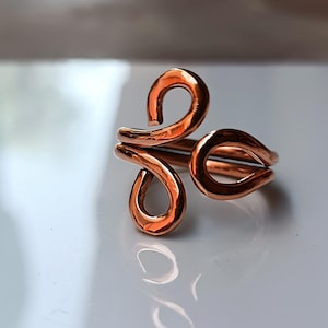 May include: A copper-colored ring with a unique, abstract design. The ring features three looped sections, creating an open, flowing shape. The metal has a polished, reflective surface, catching the light and highlighting the curves of the design.