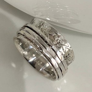 May include: A silver-colored spinner ring with a floral design. The ring features a central band with a hammered texture, surrounded by several smooth, rotating bands. The outer band is adorned with a detailed floral pattern.