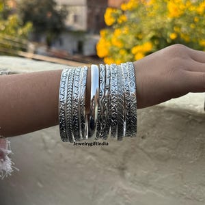 May include: A collection of silver bangles with various embossed designs, including floral and leaf patterns. The bangles are worn on a person's arm, showcasing their intricate details and polished finish. The image is taken outdoors with a blurred background.