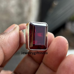 Op de afbeelding: Een zilveren ring met een grote, rechthoekige, donkerrode edelsteen. De ring wordt gedragen aan een vinger.