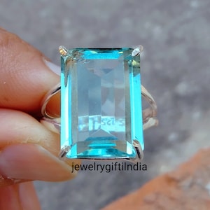Può includere: Un anello d'argento con una grande pietra rettangolare di colore blu chiaro. La pietra preziosa è sfaccettata e ha un aspetto chiaro e vitreo. L'anello è incastonato in uno stile semplice ed elegante.