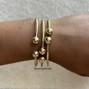 Puede incluir: Cuatro pulseras de brazalete doradas con acentos de bola redonda. Las pulseras están apiladas en la muñeca de una persona.