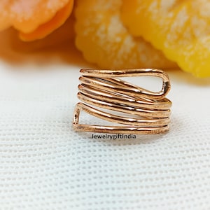Può includere: Un anello di filo color rame con un design avvolto. L'anello è composto da diversi fili di filo avvolti l'uno attorno all'altro. L'anello è su una superficie bianca con fiori arancioni e gialli sfocati sullo sfondo. Il testo "JewelrygiftIndia" è visibile.