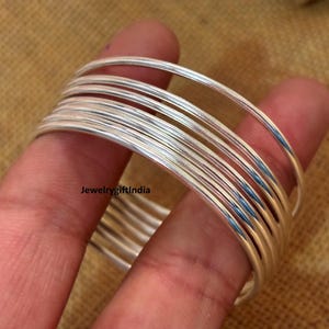 May include: A stack of silver bangles, each with a smooth, reflective surface. The bangles are stacked on a finger, with the text "Jewelrygiftindia" visible. The jewelry is set against a warm, neutral background.