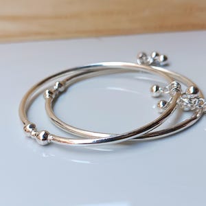 May include: Two silver bangles with small, round embellishments. One bangle has three sets of three small, dangling silver balls. The other bangle has three sets of two small, round embellishments.