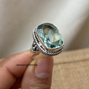 Puede incluir: Un anillo de plata con una gran gema ovalada de color verde claro. La piedra está engastada en un bisel trenzado decorativo. La banda del anillo tiene un diseño detallado. El texto "JewelrygiftIndia" es visible.