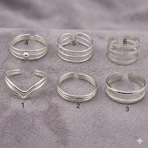 May include: A collection of six silver toe rings. Designs include a V-shape, multiple bands, and a ring with a small ball. The rings are displayed on a textured, purple surface.