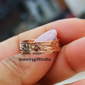 May include: A rose gold ring with a floral and leaf pattern. The ring features a continuous design of flowers and foliage around the band. The ring is held in a hand, showcasing the intricate details of the engraved design.
