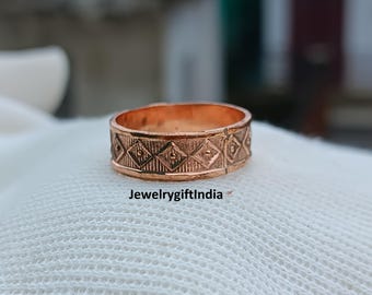May include: A copper-coloured ring with a geometric pattern of diamonds and squares. The ring is set on a white surface. The text "JewelrygiftIndia" is visible on the surface.
