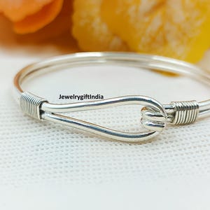 May include: A silver bangle bracelet with a hook and eye clasp. The bracelet has a wrapped wire design on either side of the clasp. The text "jewelrygiftindia" is visible on the bracelet.