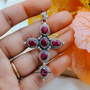 May include: A silver cross pendant featuring oval, deep red gemstones. The pendant has an ornate design and the text "jewelrygiftindia". The cross is held in a hand.