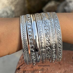 May include: A set of silver bangles with intricate designs, including hearts, flowers, and geometric patterns. The bangles are stacked on a person's wrist.