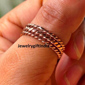 Op de afbeelding: Close-up van drie koperkleurige, gedraaide ringen in touwstijl, gestapeld op een vinger. De ringen hebben een textuur en een warme, metallic glans. De ringen zijn van Jewelrygiftindia.