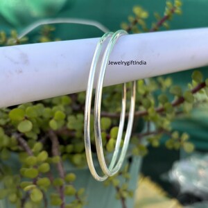 May include: Two silver bangles, one slightly larger than the other, are stacked on top of each other. The bangles are smooth and shiny, and they are resting on a white surface.