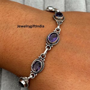 May include: A silver bracelet with oval purple gemstones. The bracelet has a delicate design with a rope-like border around each stone. The text "JewelrygiftIndia" is visible on the image. This piece of jewellery is a bracelet.