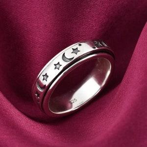 May include: A silver-colored ring with a celestial design featuring stars and crescent moons. The ring has a rotating outer band and is set against a deep red background. The ring is marked with "925".