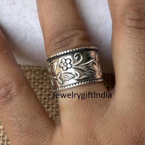 Peut inclure: Une bague en argent avec un motif floral. La bague est large et a une surface texturée. La bague est portée au doigt d'une personne.