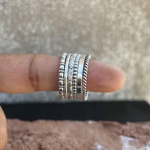 May include: A set of five silver stacking rings with different textures and patterns. The rings are worn on a finger.